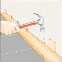 Drawing of a man's hand, nailing a floor board beside a wall base spacer.
