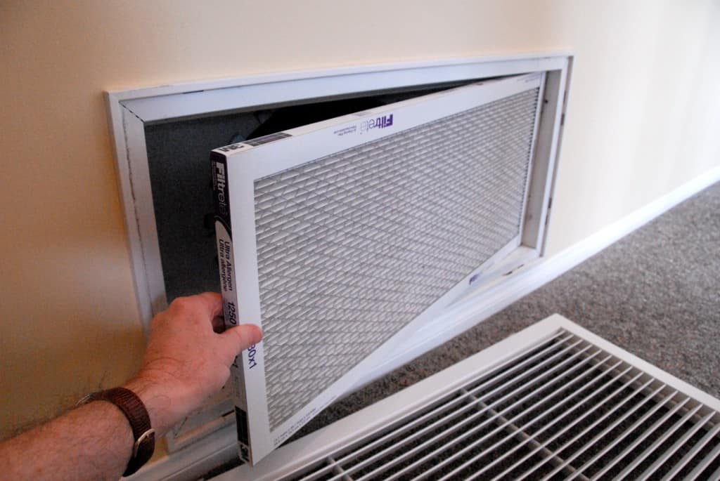 A hand placing a new air filter in a wall register, with airflow arrows pointing toward the ductwork.