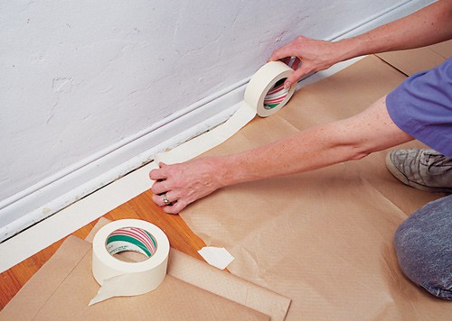 Woman's hands, securing a drop cloth along baseboards with a wide masking tape.