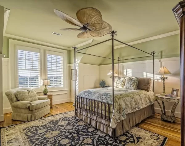 Vintage table lamps, a decorative ceiling fan, an ornate canopy bed, a decorative rug, and an overstuffed chair give this bedroom luxurious style.